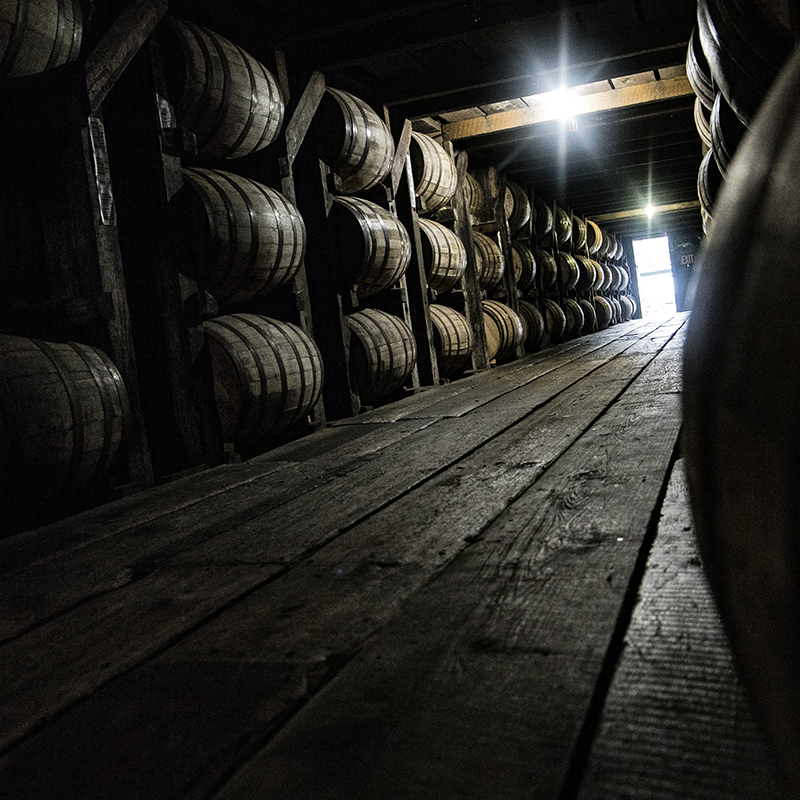 Dimly lit wooden warehouse interior with rows of stacked barrels on both sides, leading to a bright doorway at the end.