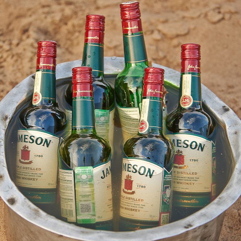 Six Jameson whiskey bottles in a metal bucket filled with water, placed on a sandy surface.