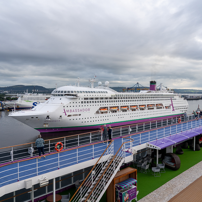 Ambience docked at a port, with a cloudy sky and distant hills in the background.