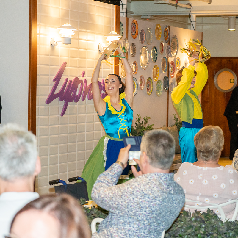 Dancers in colorful costumes perform energetically on Lupino's Stage, with a decorative wall and audience in the foreground.