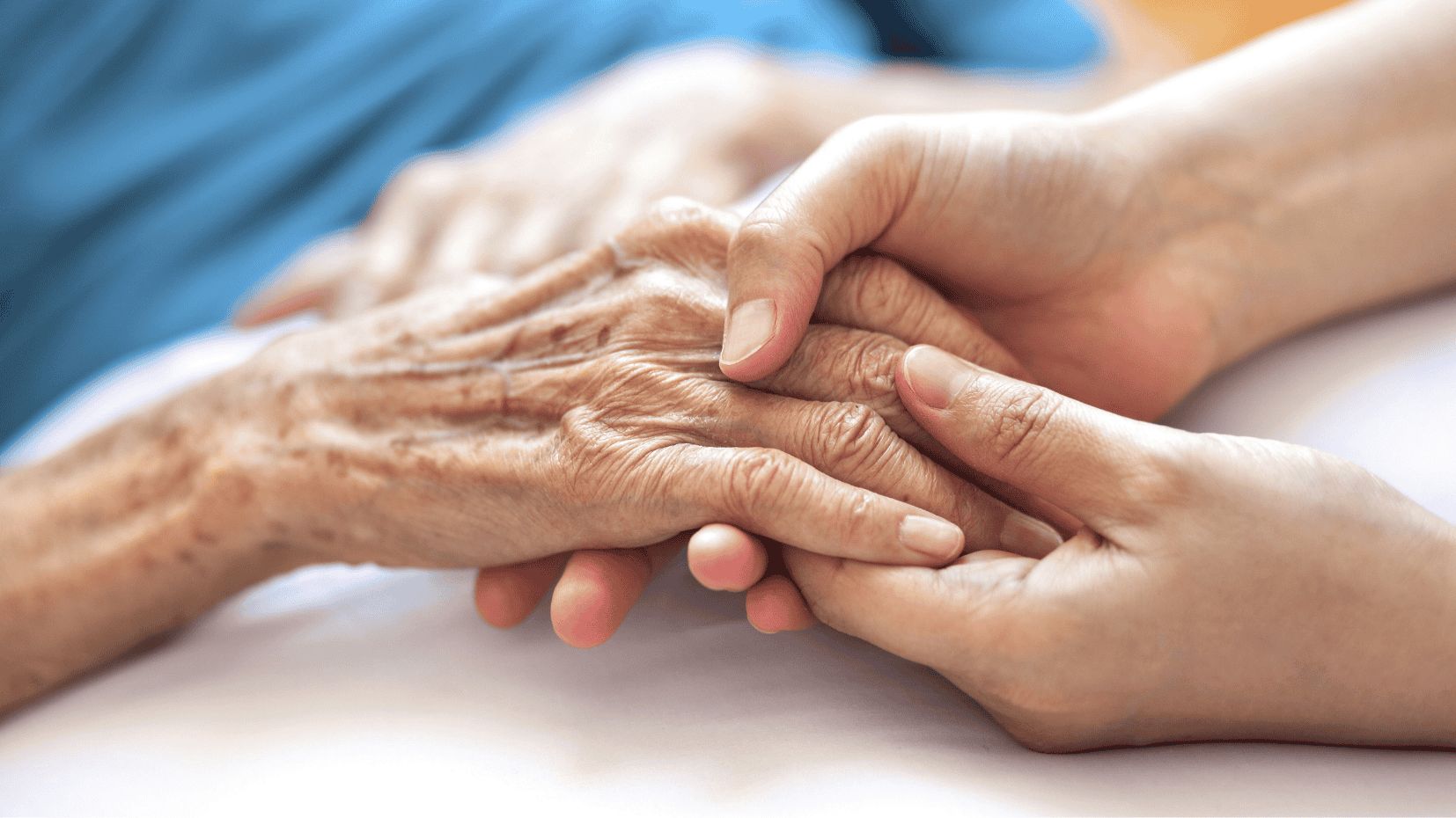 A younger person gently holds the hand of an elderly person, conveying care and support, with a soft blue background.