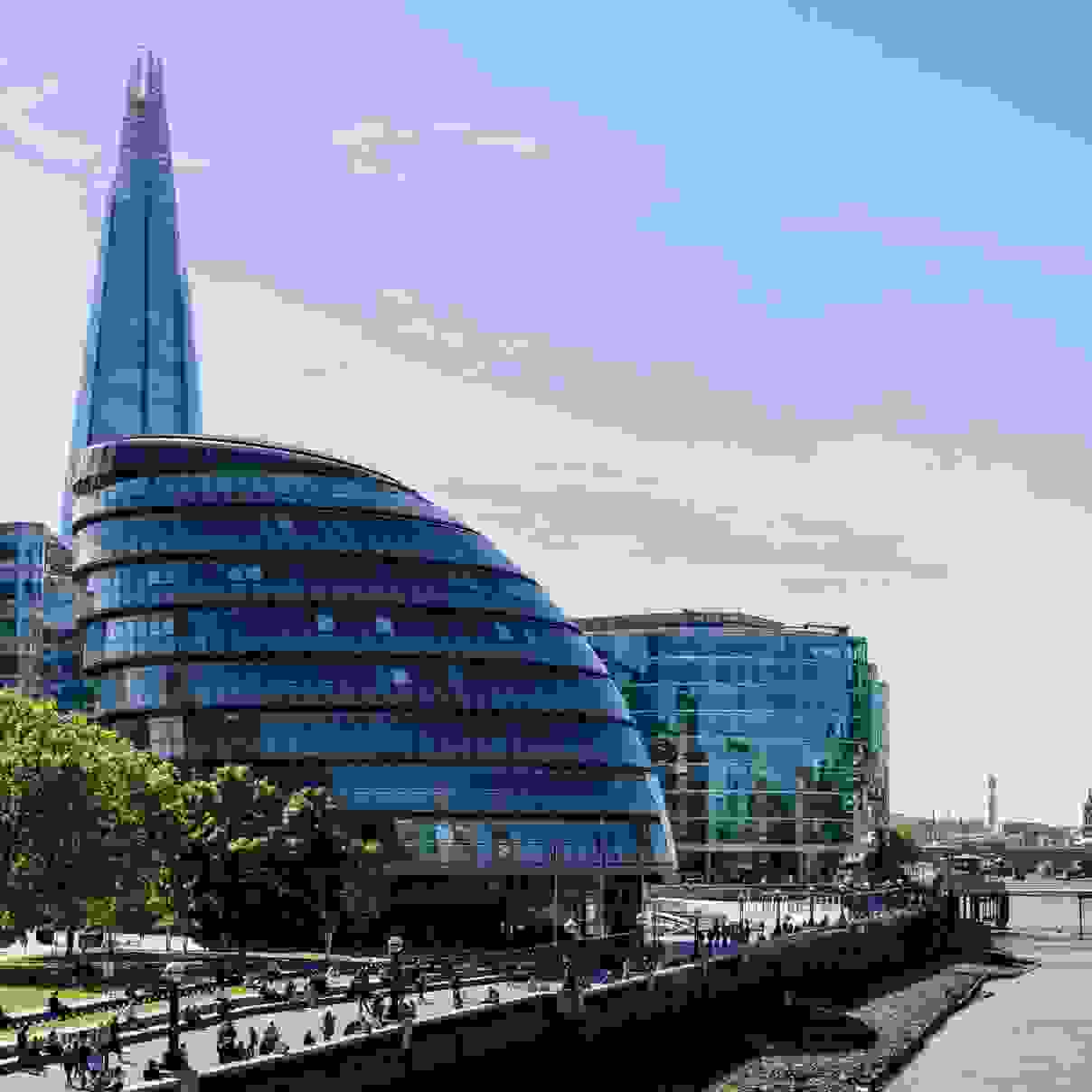 A scenic view of london's skyline featuring the shard and city hall beside the thames river on a sunny day, with people walking along the riverbank.