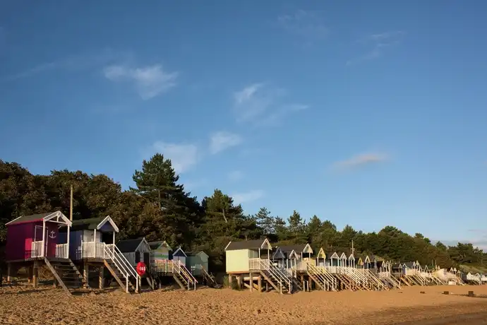 Colorful beach huts on wooden stilts line a sandy shore, with trees and a clear blue sky in the background.