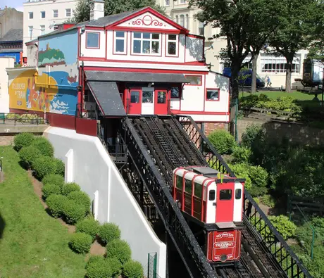 A red and white funicular tramway midway up the cliff in Scarborough, with green trees and the top station showing