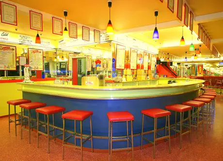 Retro-style ice cream bar and diner with a curved counter, red stools, colorful hanging lights, and menu boards in a vibrant, well-lit setting.