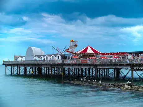 A seaside pier with a white and red striped tent, amusement rides, and people walking. Seagulls rest on rocks below under a cloudy blue sky.
