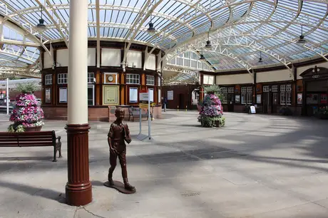 Spacious train station with ornate glass ceiling, a bronze statue of a boy, benches, and vibrant flower arrangements.