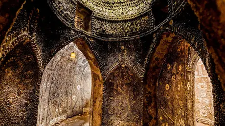 Interior of a grotto with intricate shell mosaics covering walls and arches, illuminated by warm lighting.