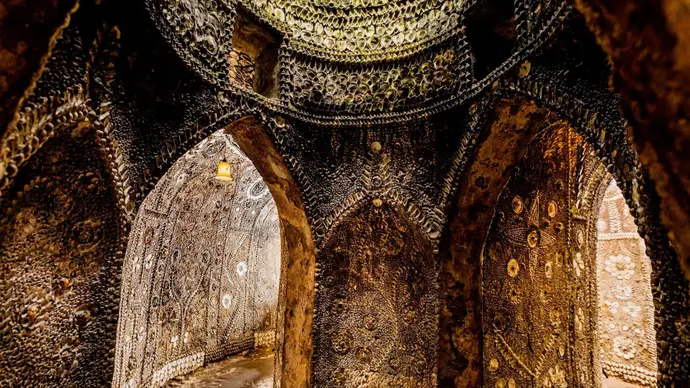 Interior of a grotto with intricate shell mosaics covering walls and arches, illuminated by warm lighting.