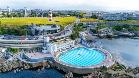 Aerial view of Plymouth Hoe, featuring a lighthouse, wide green space, and Tinside Lido pool, with the cityscape in the background.
