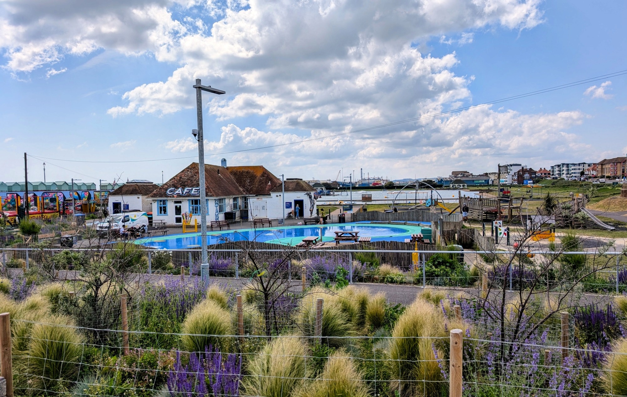 Coastal scene with a café, the Lagoon, and colorful play area surrounded by lush greenery and purple flowers under a partly cloudy sky.
