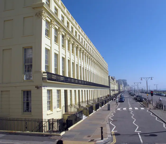A view along Hove promenade, showing Georgian buildings on the left, and the sea and beach on the right, under a blue sky.