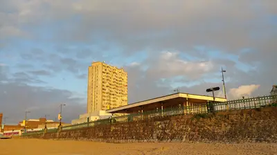 Tall apartment building and seafront promenade under a cloudy sky, illuminated by warm sunlight, with sandy beach in the foreground.