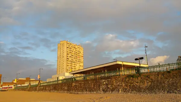 Tall apartment building and seafront promenade under a cloudy sky, illuminated by warm sunlight, with sandy beach in the foreground.