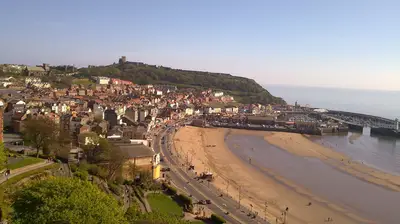 Scarborough south bay with a sandy beach, colorful buildings, and a harbor. A castle on a hill overlooks the town, set against a clear blue sky and calm sea.