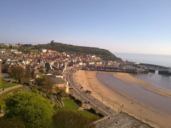 Scarborough south bay with a sandy beach, colorful buildings, and a harbor. A castle on a hill overlooks the town, set against a clear blue sky and calm sea.