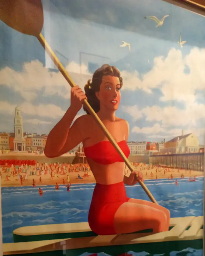 A vintage railway poster of Herne Bay showing a woman in a red bikini on a paddle board
