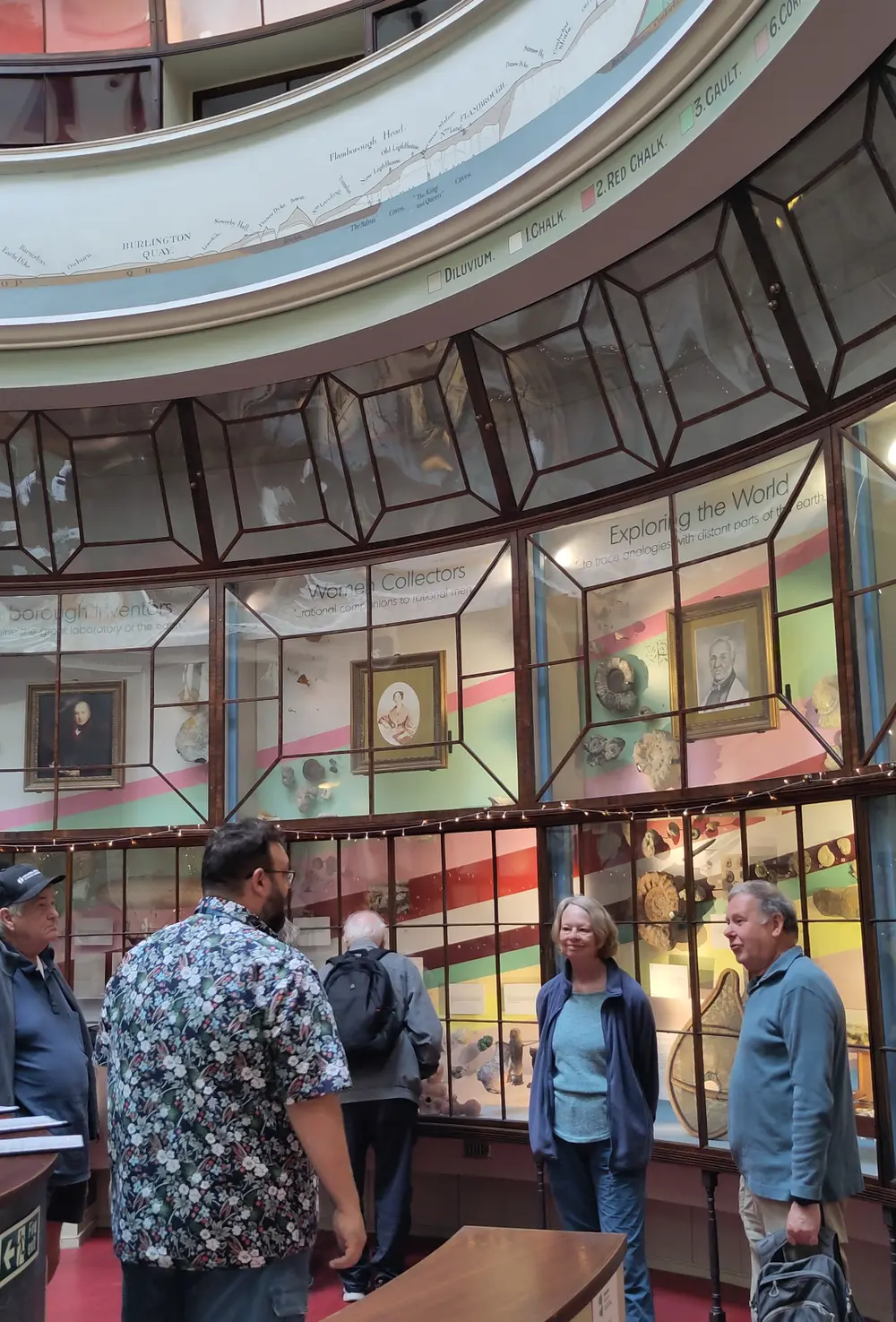 People exploring a museum exhibit with displays and portraits on the walls, featuring geological and historical themes.