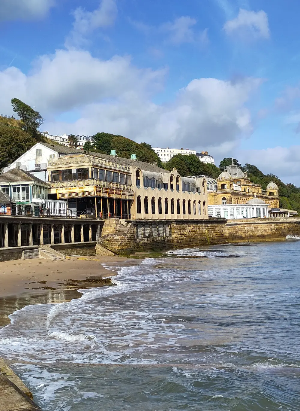 A seaside view with waves washing onto a sandy shore, historic buildings on the cliffside, and a bright blue sky with fluffy clouds.