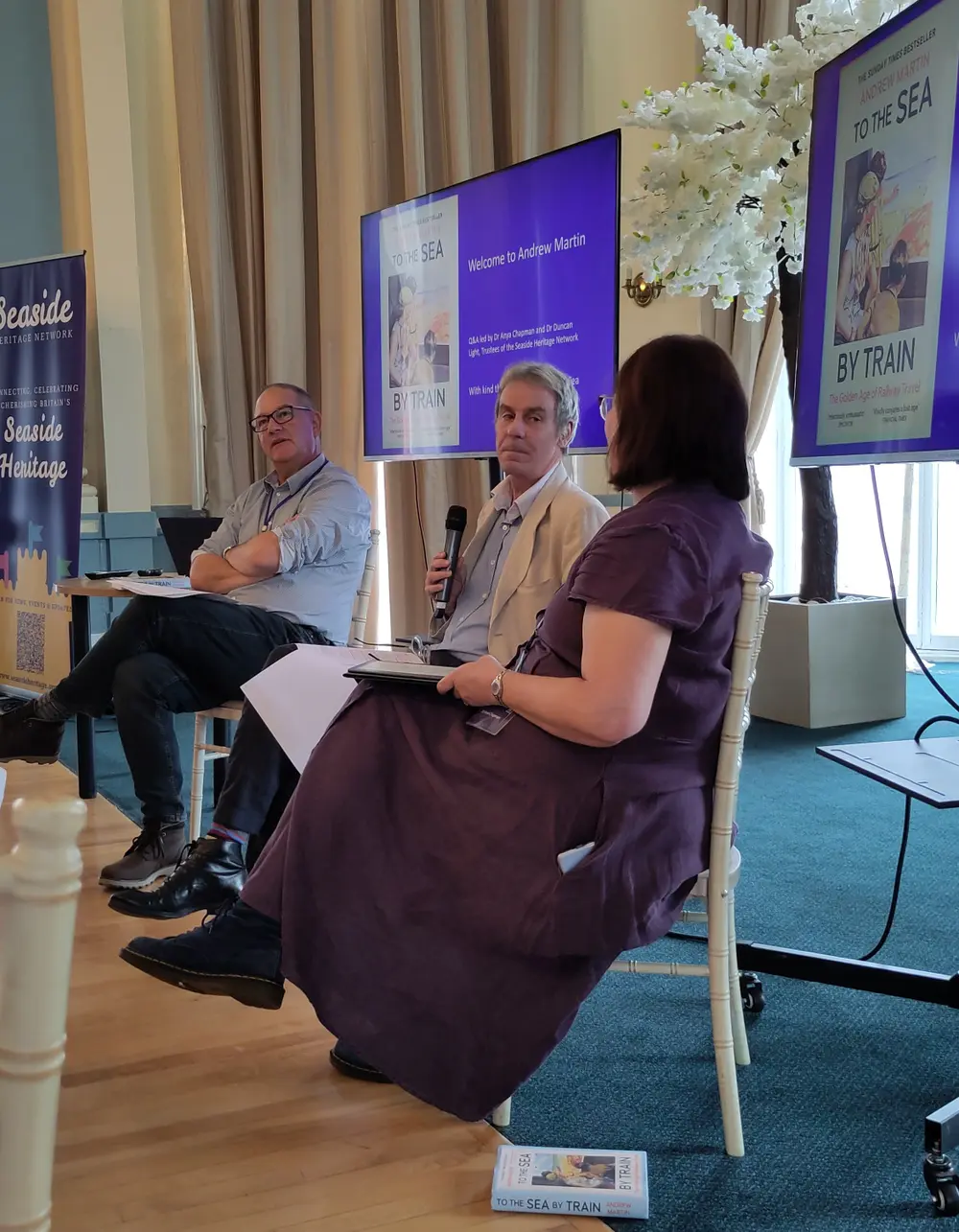 Three people seated onstage during a discussion, with a microphone, books, and a presentation screen titled "To the Sea by Train" in the background.
