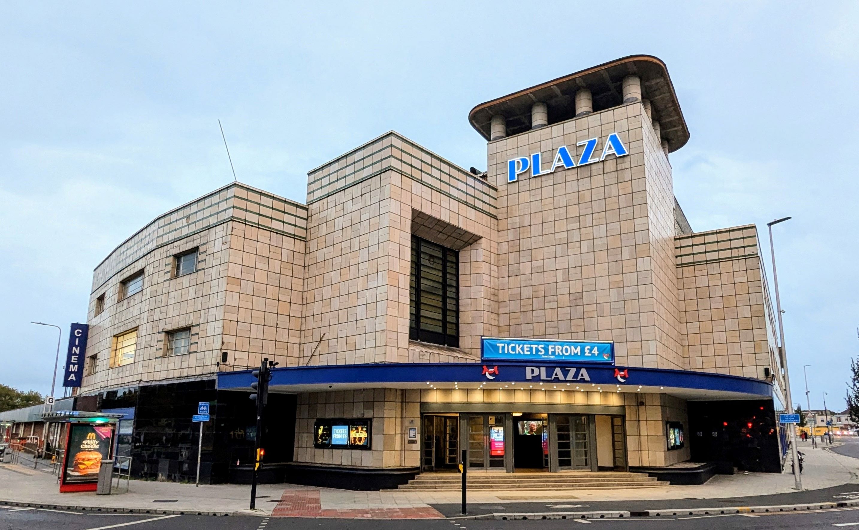 Art Deco-style cinema building with beige tiles, blue "Plaza" sign, and ticket booth. Posters and lights adorn the entrance.