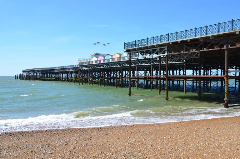 A historic pier extends over the sea, with multicolored beach huts on top and gentle waves lapping against the pebbled shore under a clear blue sky.
