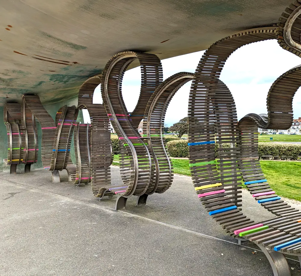 A modern sculpture with curving wooden slats and colorful accents under a concrete roof on a paved area with grass and bushes nearby.