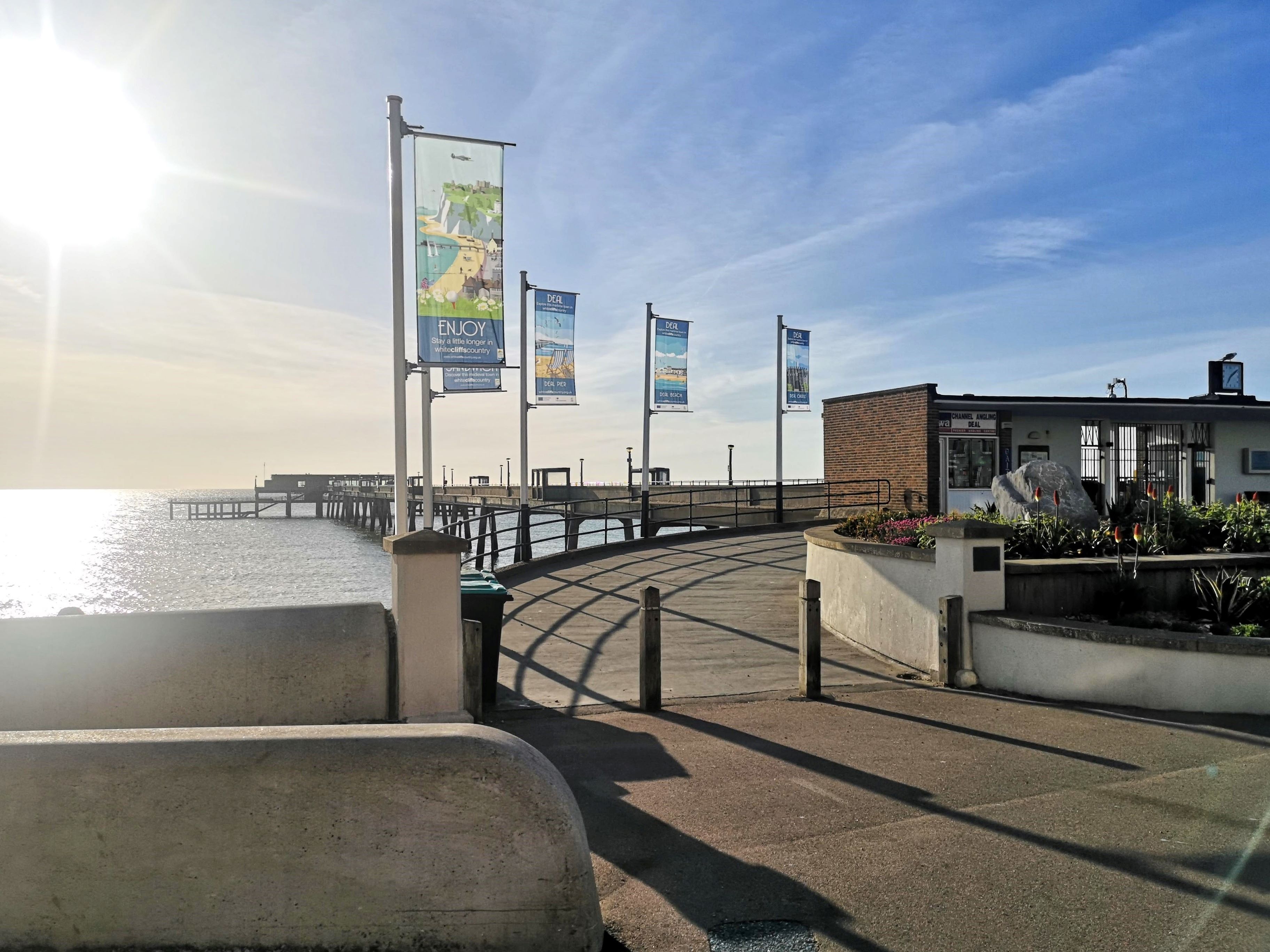 A sunny seaside pier with colorful banners, a building to the right, and a clear blue sky reflecting on the calm water.