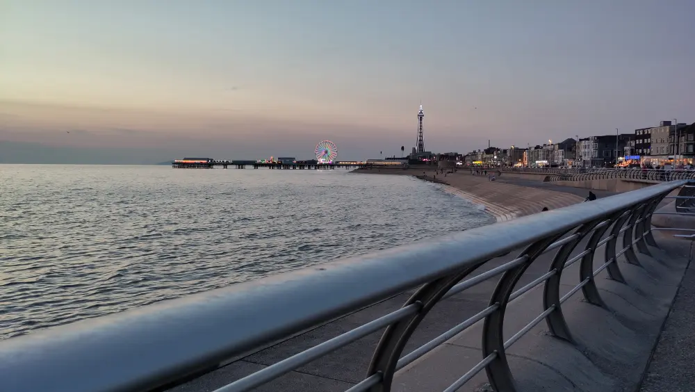 Promenade at dusk with a Ferris wheel and tower in the distance, lights reflecting on calm sea, railing in foreground, and buildings lining the coast.