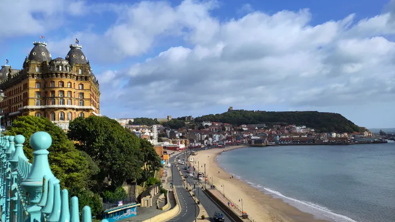 The Grand Hotel overlooking a sandy beach and sea, with Scarborough and green hills in the background under a partly cloudy sky.