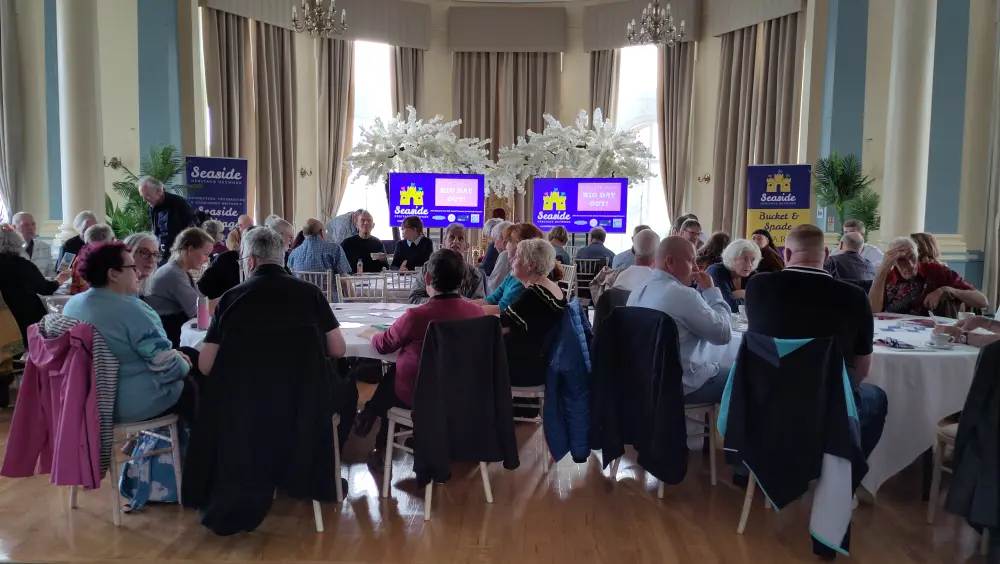 A group of people seated around tables at a conference in a large room with decorative plants and banners reading "Seaside Heritage Network" and "Big Day Out".