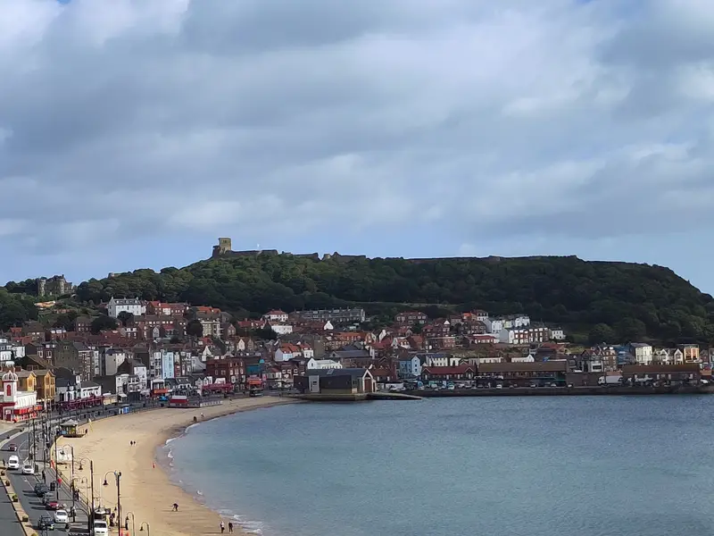 Coastal town view with a grand historic hotel, sandy beach, winding road, and hillside houses under a partly cloudy blue sky.
