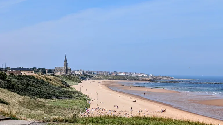 A beach under blue skies with people sunbathing and paddling at the resort of Tynemouth