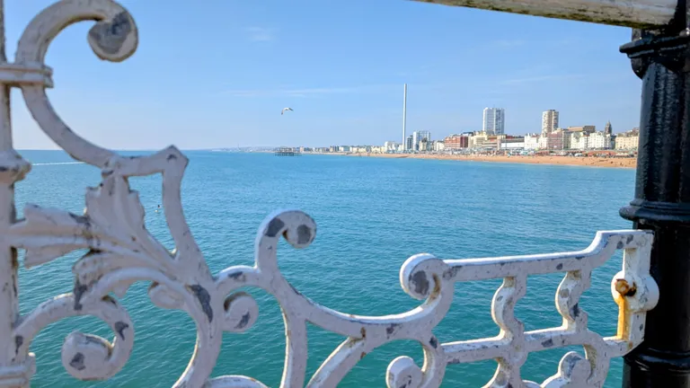 View of Brighton seafront through ornate white railing, featuring blue ocean, a beach, buildings, and a distant pier under clear blue skies.