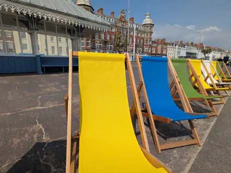 Colorful deck chairs in yellow, blue, and green line a seaside promenade with historic buildings and a pavilion in the background.