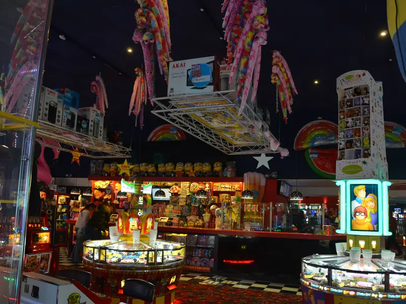 Brightly lit arcade with colorful stuffed animals, hanging decorations, gaming machines, and prize displays. People are playing games.