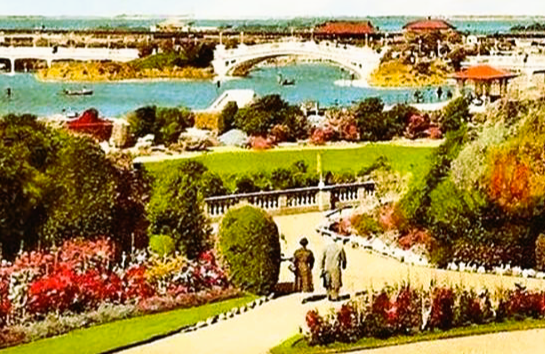 A colorful garden overlooks a lake with elegant bridges, surrounded by lush greenery and vibrant flowers. Two people walk along the curved path.