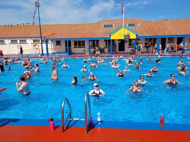 People enjoying a sunny day in a large outdoor swimming pool, with a colorful building and flags in the background.