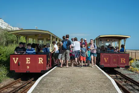 Crowd boarding a vintage open-air train at a seaside station, with blue sky and greenery in the background.