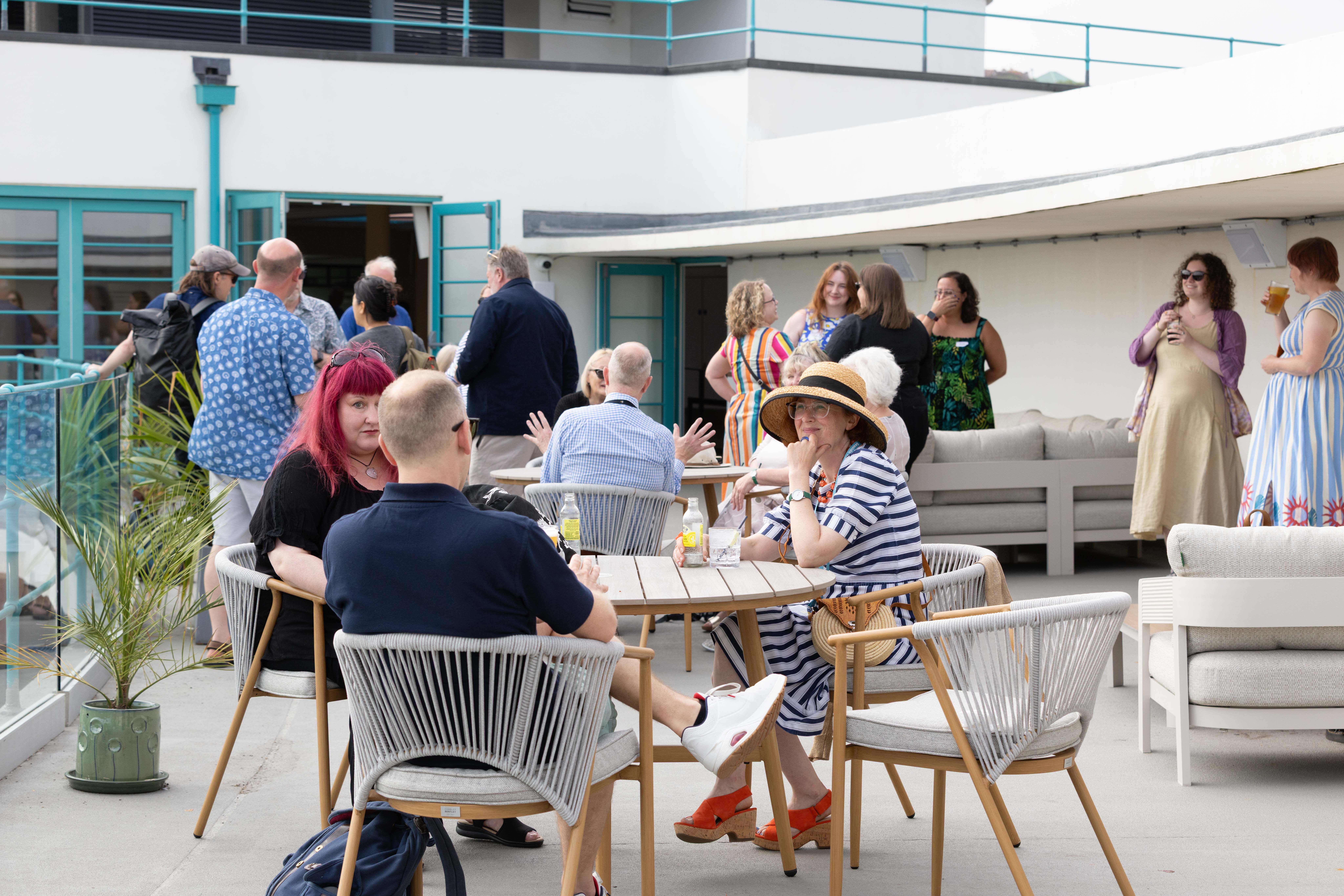 People socializing and sitting at tables on a rooftop terrace. Some are standing, while others are seated, enjoying drinks and conversation.