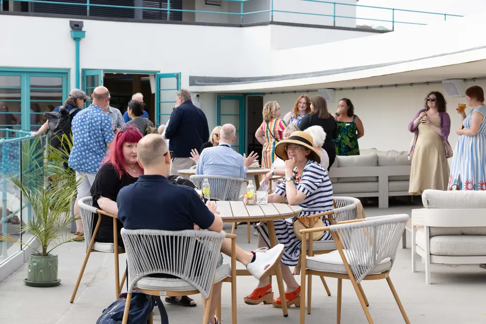 People socializing and sitting at tables on a rooftop terrace. Some are standing, while others are seated, enjoying drinks and conversation.