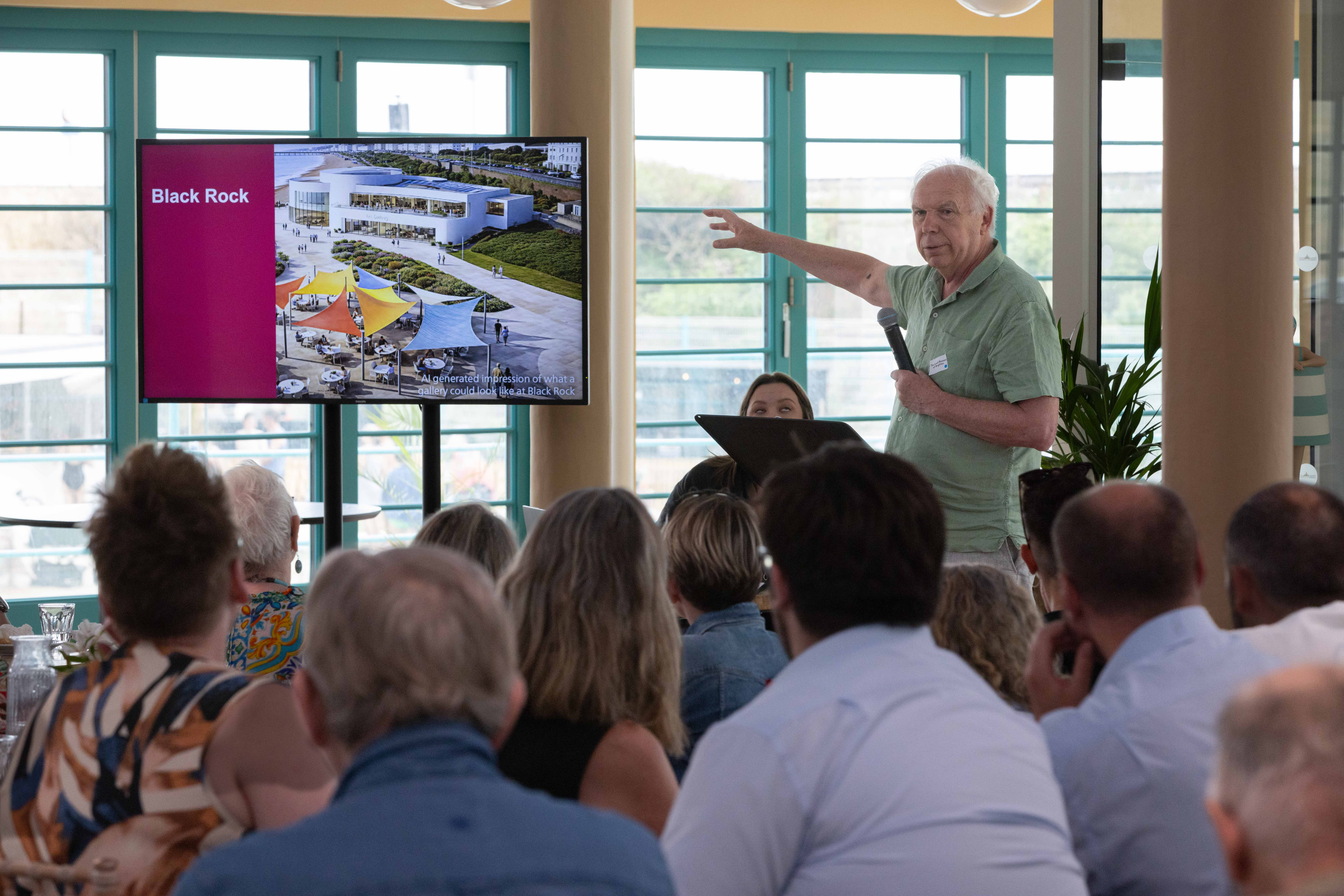 A man in a green shirt presents to an audience, pointing at a screen displaying a gallery design labeled "Black Rock" with colourful canopies.