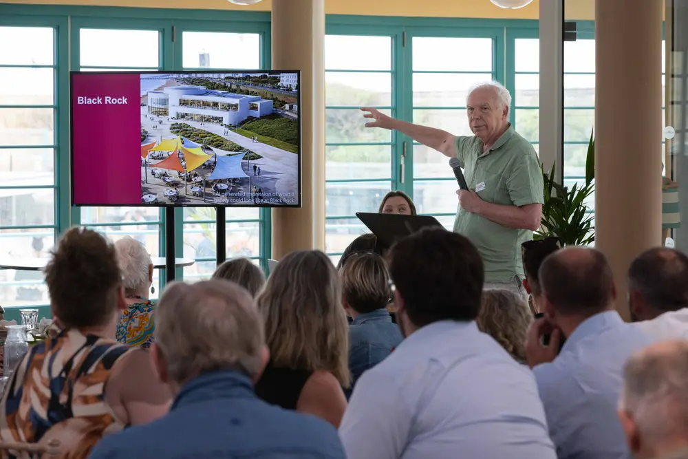 A man in a green shirt presents to an audience, pointing at a screen displaying a gallery design labeled "Black Rock" with colourful canopies.