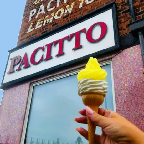 Hand holding a lemon-topped ice cream cone in front of Pacitto's shop with a brick facade and vintage signage.
