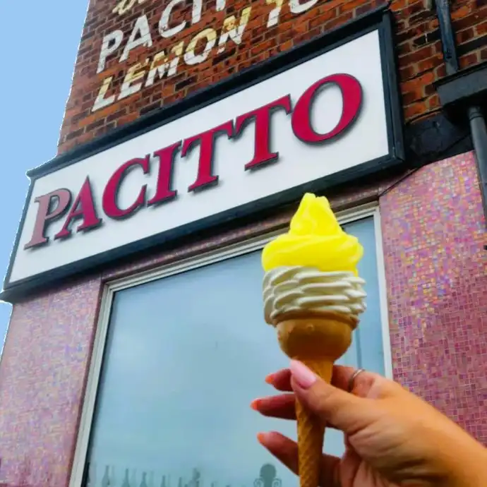 Hand holding a lemon-topped ice cream cone in front of Pacitto's shop with a brick facade and vintage signage.