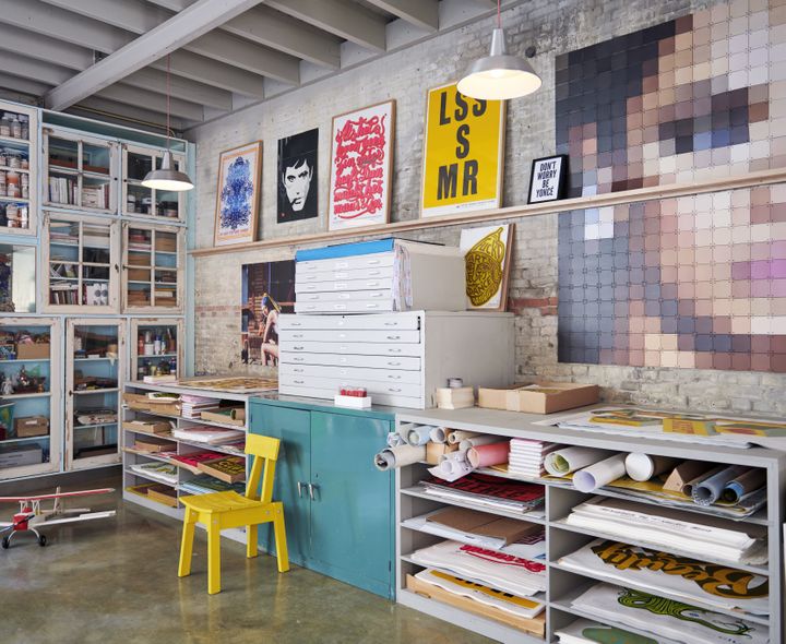 Een bakstenen muur met ingelijste posters, een grote pixel-IXXI en werkbanken vol prints