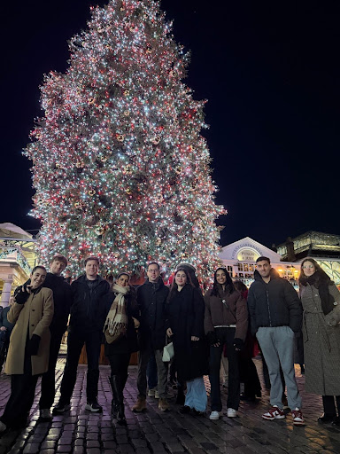 ARK members posing in front of a large, brightly lit Christmas tree at night in a cobblestone plaza.