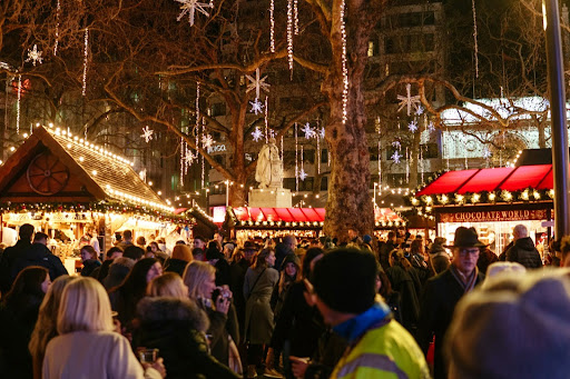 Crowded outdoor Christmas market at night with twinkling lights, hanging snowflake decorations, wooden stalls and visitors in winter coats.