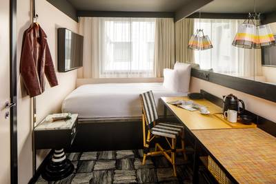 Compact hotel room with a bed, desk, chair, and wall-mounted TV. Jacket hangs by the door, and colorful lampshades adorn the ceiling.