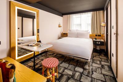 Modern hotel room with a large bed, decorative stool, marble-top table, and illuminated mirror. Neutral tones and geometric carpet design.